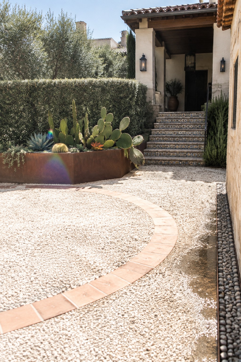 Curved gravel courtyard with rusty succulent planter and colorful tiled staircase at villa entrance
