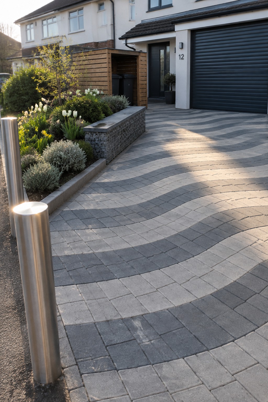 A small modern driveway paved with gray blocks in a wavy curved pattern, bordered by a gabion retaining wall and garden bed with shrubs and flowers.
