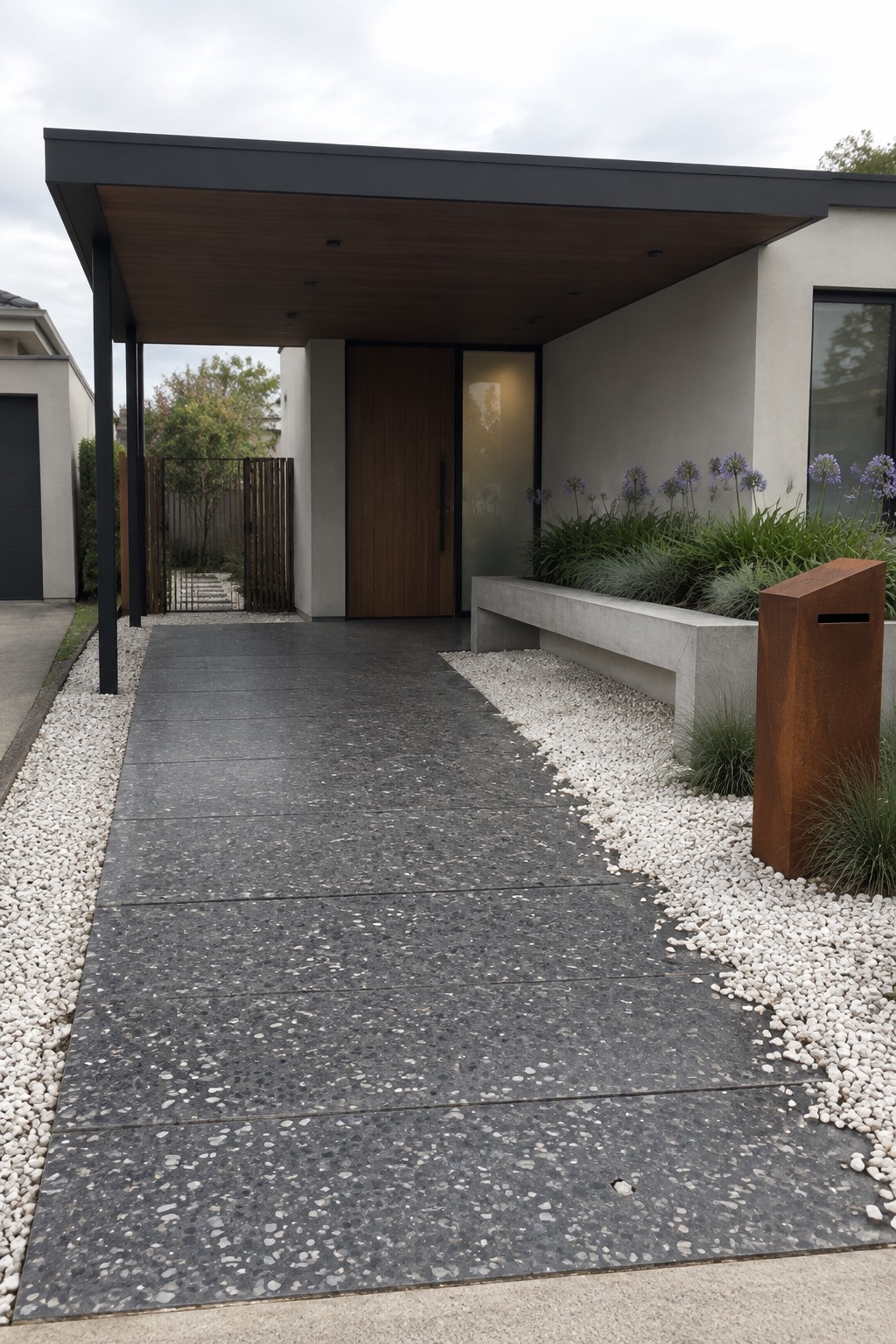 A modern house entrance featuring a dark speckled concrete paver driveway bordered by white gravel, leading to a wooden door beneath a covered porch with a concrete bench and rusted metal post on the right.