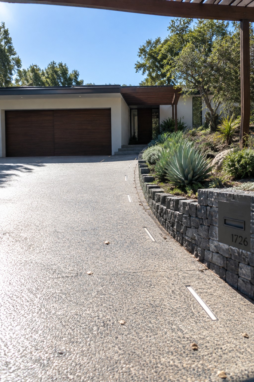 A wide concrete driveway with embedded linear lights leads to a low-profile modern house featuring a dark wood garage door and a stone retaining wall with landscaping on the right side.