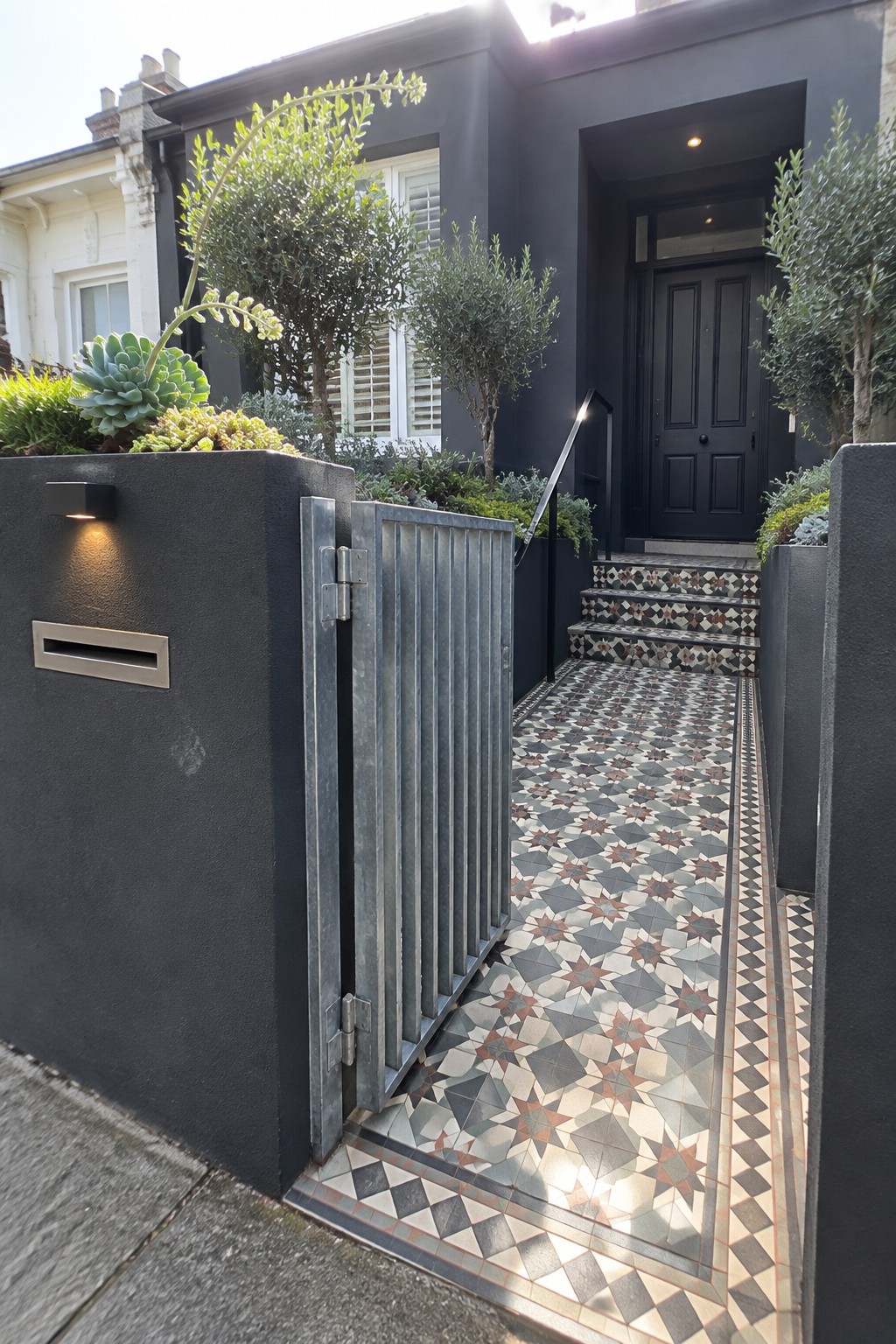 A metal vertical slat gate is set into a dark boundary wall beside a patterned tile path leading to steps and a black front door.