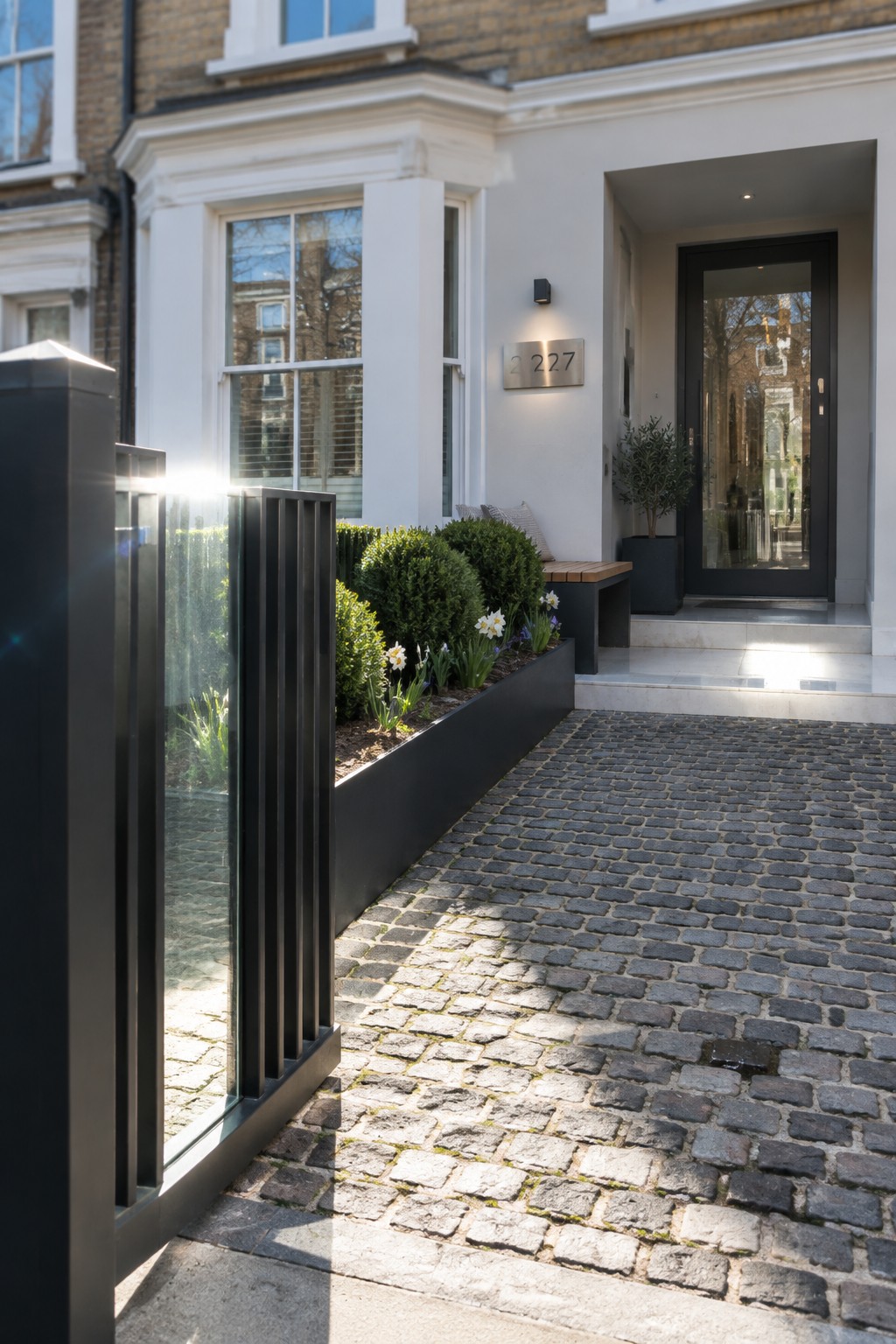 A black metal vertical slat fence with a glass panel runs beside a cobblestone driveway, next to a long raised black planter filled with shrubs and flowers, in front of a white house entrance.