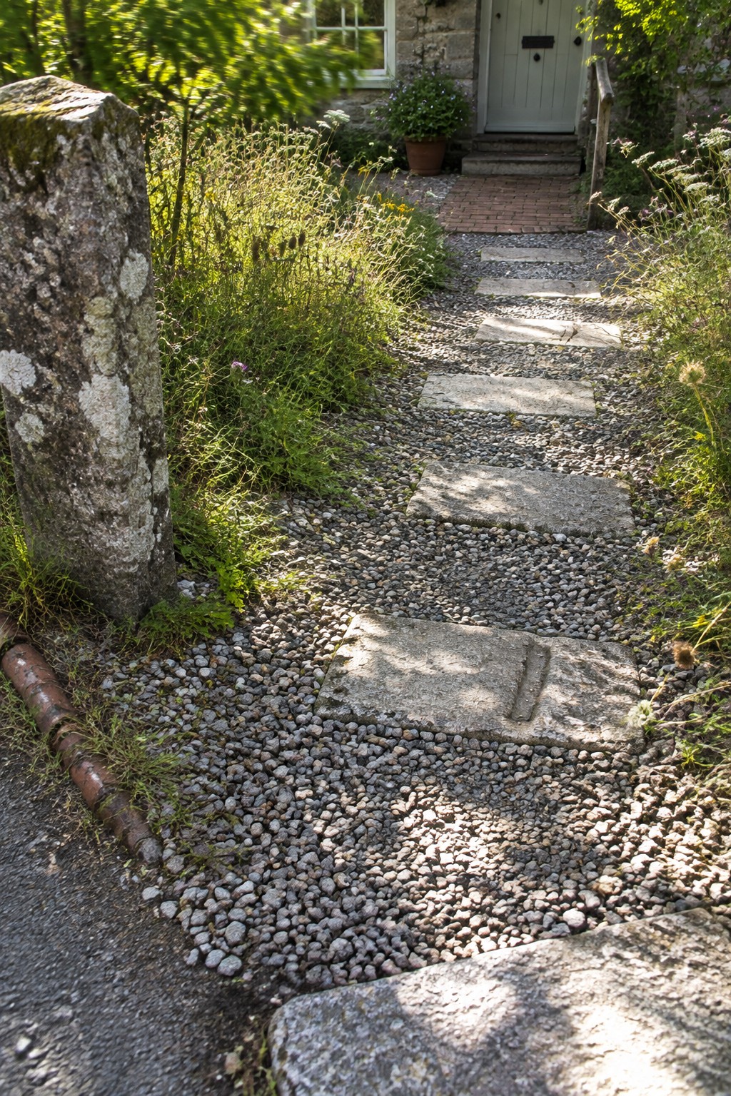 A gravel path lined with rectangular stone slabs leads toward a light green door on a stone cottage, with wildflowers and greenery along both sides.