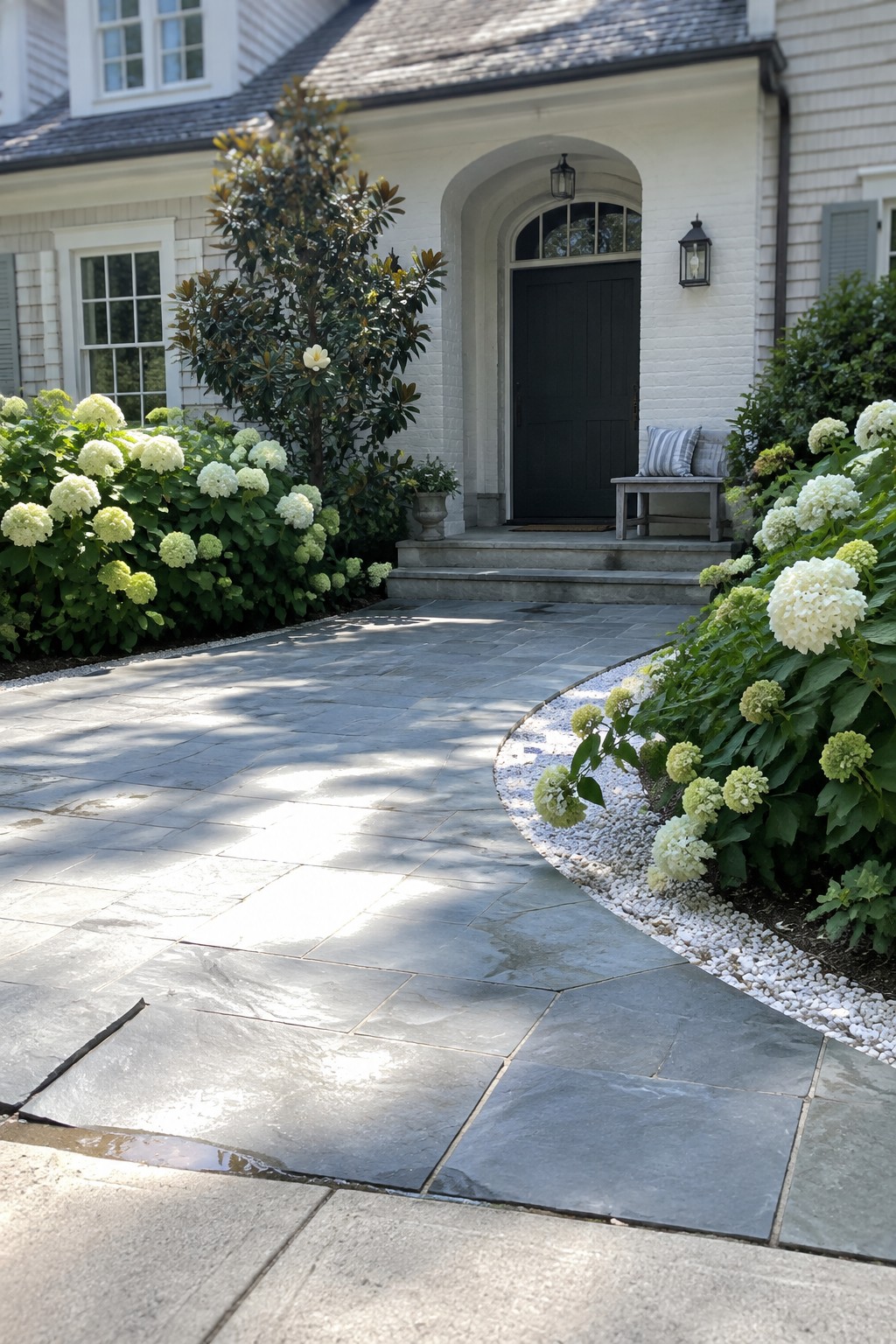 A curved gray stone driveway with white gravel edging runs past white hydrangea beds toward a white brick house with a dark arched door.