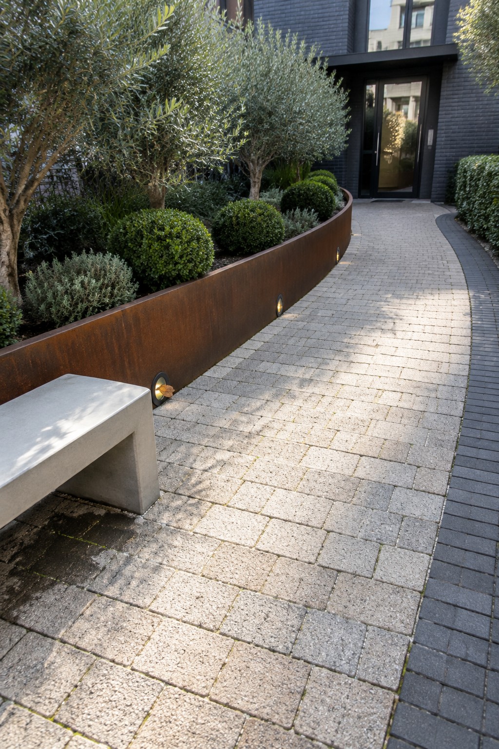 A curved paved pathway with a rusted metal border separating it from planted beds and trees next to a building entrance.