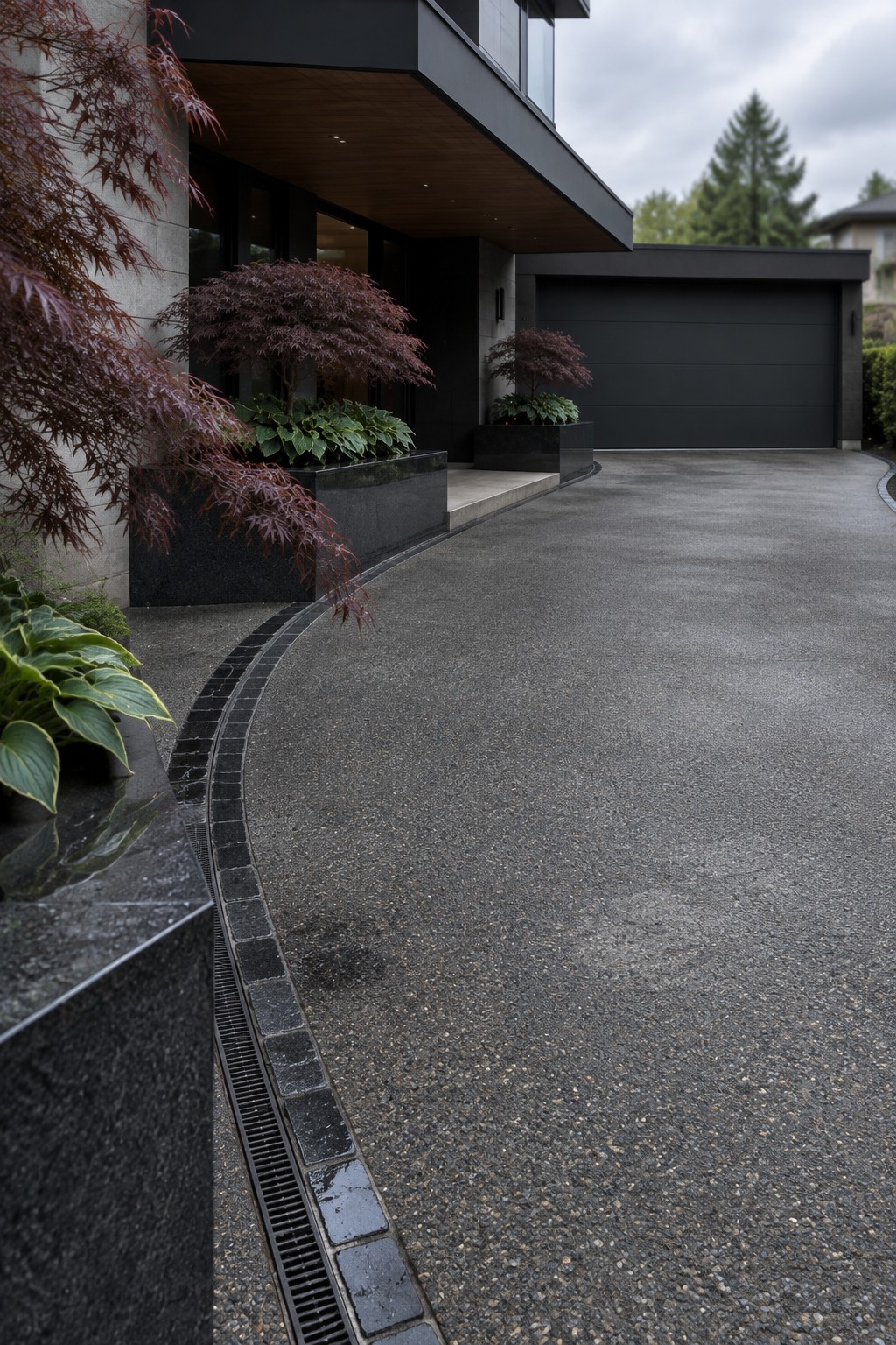 Modern house exterior showing a curved asphalt driveway with a linear drain grate and stone paver border running along the edge, flanked by dark stone planters with red foliage.