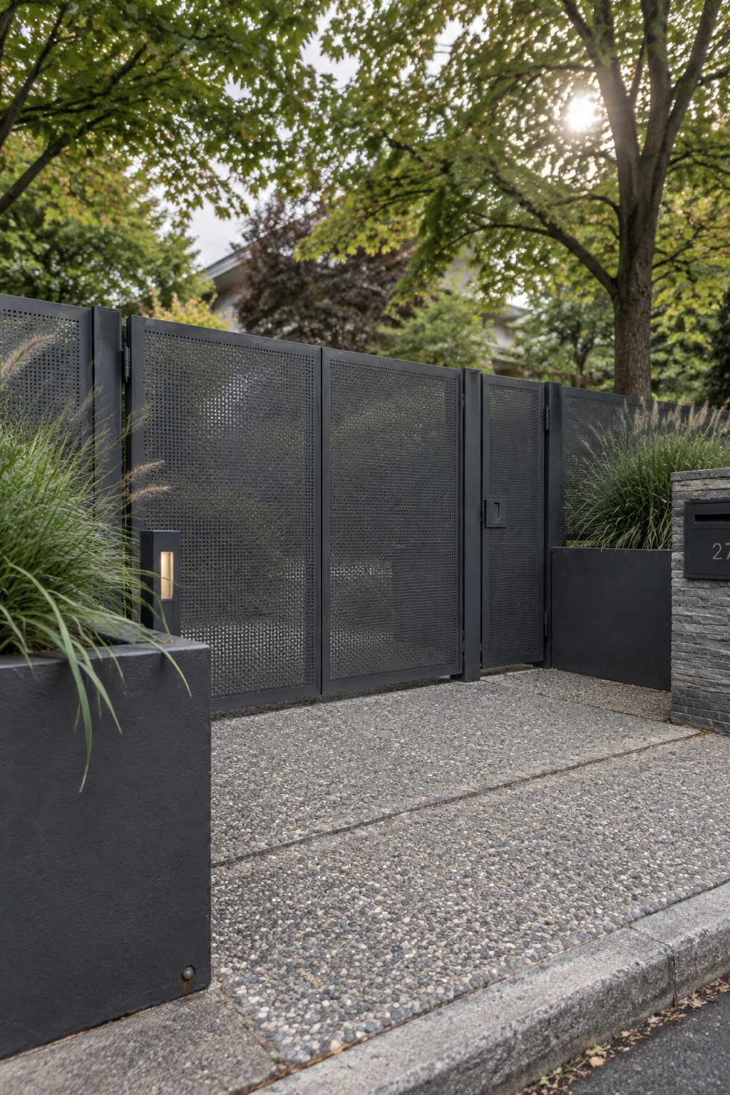 A wide black perforated metal driveway gate stands open on a gravel surface, with dark planters holding tall grasses on both sides.