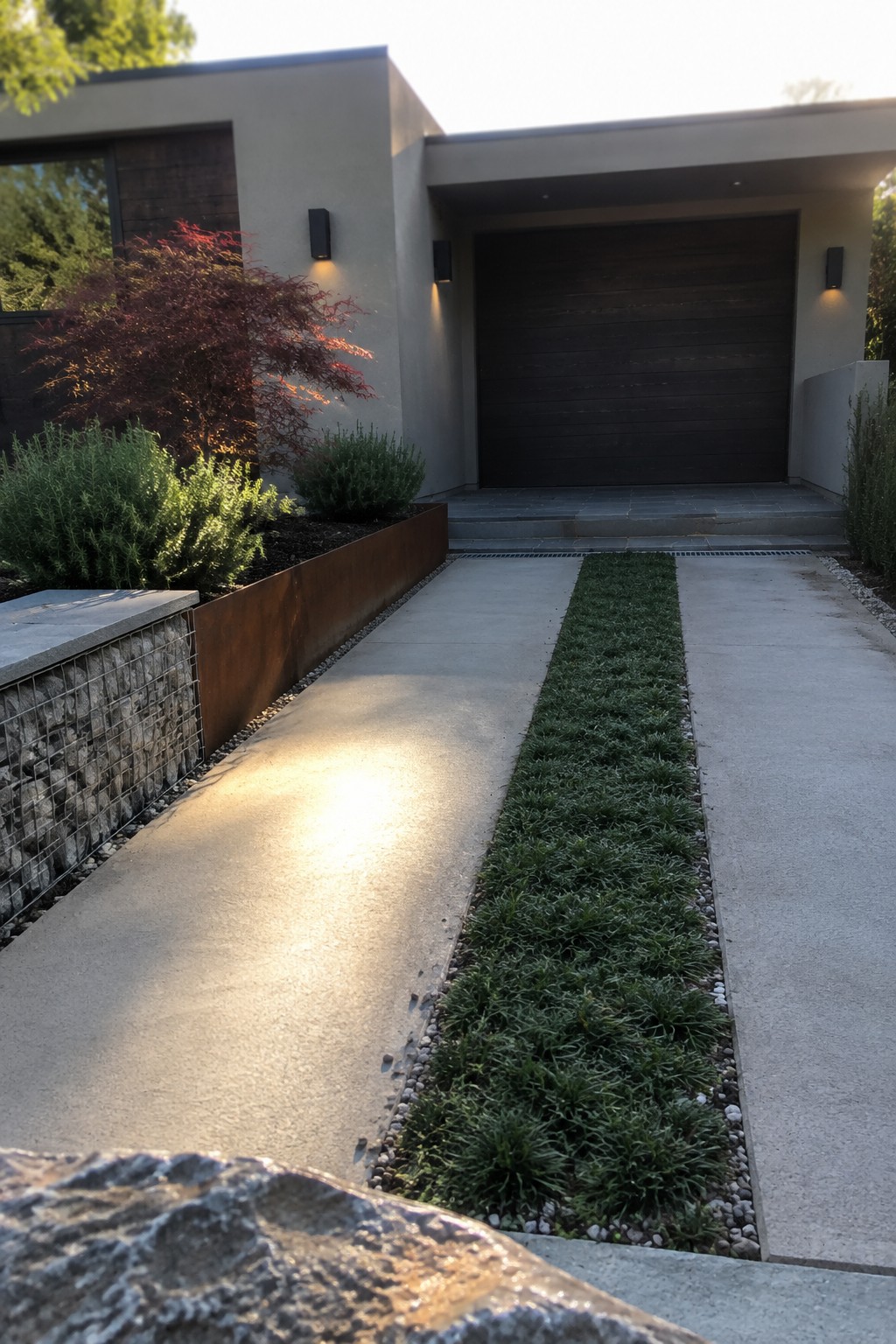 A modern concrete driveway with a central strip of grass leads to a dark wood garage door on a contemporary house, with planted beds and a rusted metal edge on the left.