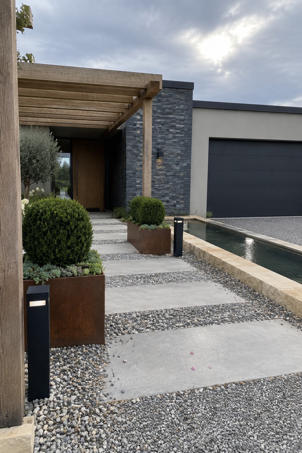A modern house entrance with concrete pavers set in gravel, large rusted metal planters holding shrubs, a narrow water feature, and a wooden pergola over the front door.