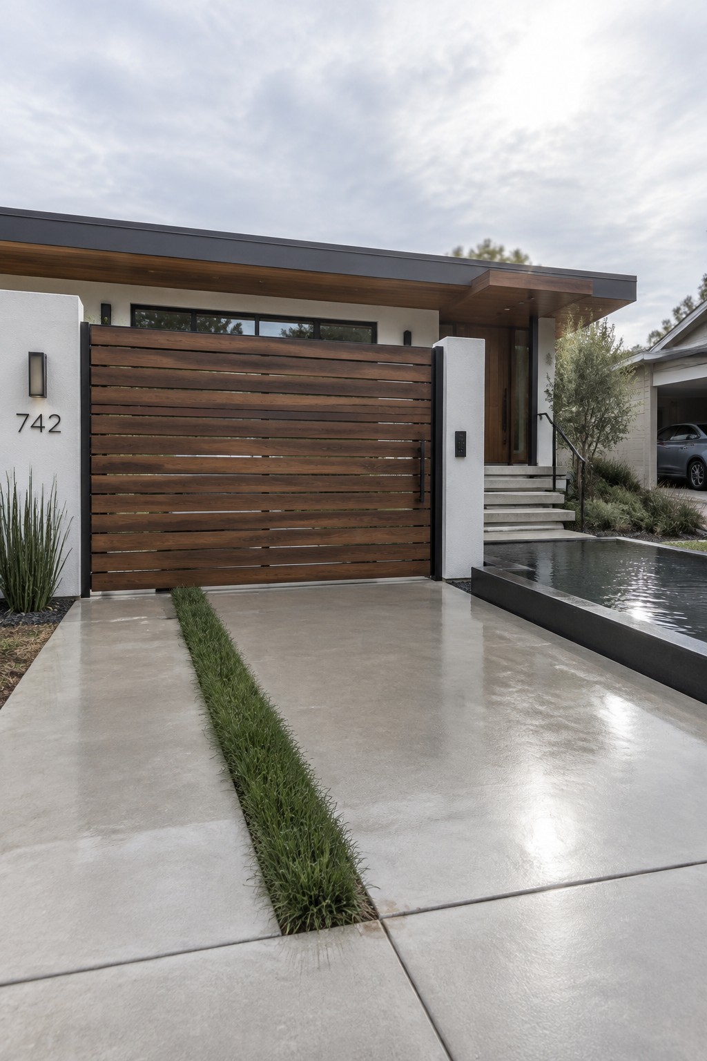 A modern house exterior featuring a large horizontal wooden slat driveway gate, concrete paving with a central grass strip, and a narrow water feature beside the entrance steps.