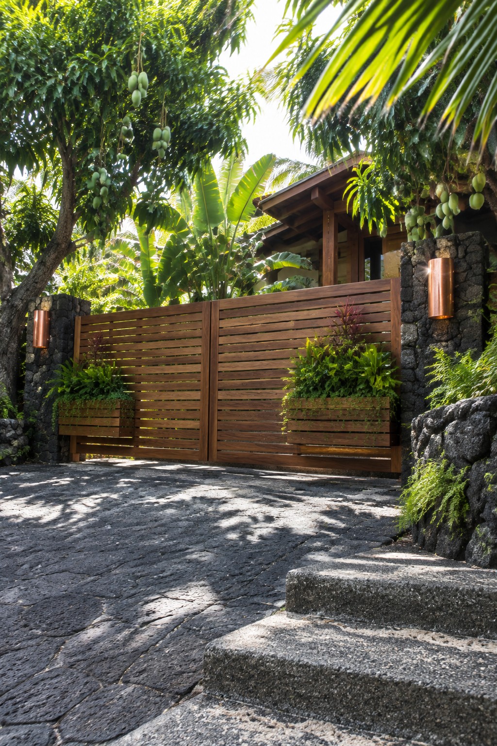A wooden driveway gate with horizontal slats set between dark stone pillars, each pillar fitted with a wooden planter box holding green plants.