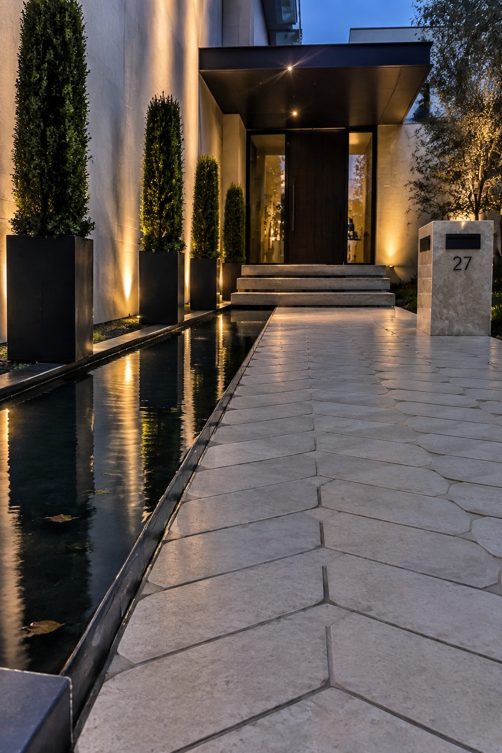 Modern home entrance with light irregular paver walkway leading to steps, narrow water channel on the left, and tall planters in black pots beside a dark wood door.